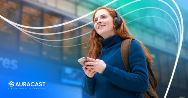 A young woman with red hair joyfully walks through an airport wearing a blue sweater and wireless headphones. The Auracast wordmark is visible in the lower left.