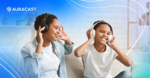 Two girls joyfully share music experiences with Auracast headphones on a cozy sofa.