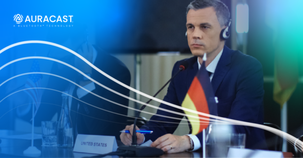 A man sitting at a conference table wearing headphones and holding a pen in front of a microphone stand and a nameplate. A logo reading Auracast, a Bluetooth technology, is visible in the upper left.