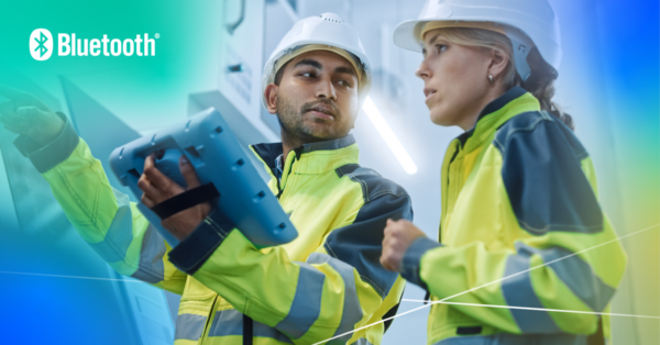 Two construction workers in high-visibility jackets and hard hats, using a Bluetooth-enabled device on a construction site.