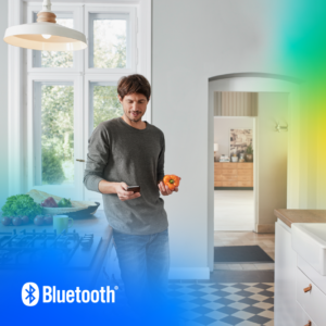 A person standing in a modern kitchen, holding an orange bell pepper in one hand and a smartphone in the other, using Bluetooth Find My to locate another device.