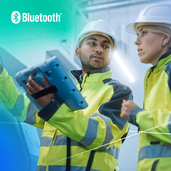 Two construction workers in high-visibility jackets and hard hats, using a Bluetooth-enabled device on a construction site.