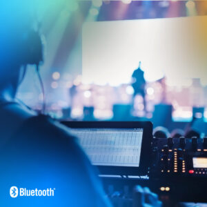 A person operates a sound mixing console at a live event with stage lights in the background, featuring the Bluetooth wordmark on the lower left corner.