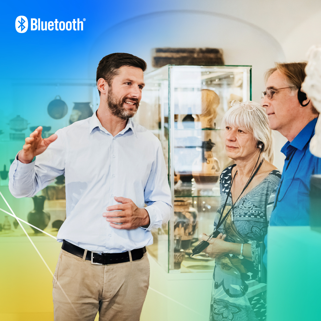 A tour guide tells the story of the surrounding art displayed in glass cases, as a man and woman listen using headphones and Auracast assistive listening technology.